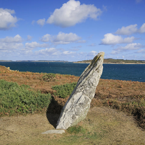 Photo from Friendly Guides Isles of Scilly Guidebook showing the Old Man of Gugh prehistoric menhir or standing stone on Gugh, St Agnes, Isles of Scilly. In the background the Garrison on St Mary's and Tresco.