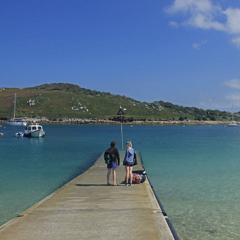 Photo from Friendly Guides Isles of Scilly Guidebook showing people waiting for the ferry on Anneka's Quay, Bryher, Isles of Scilly. In the background is Tresco.